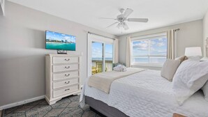Quarto com cama, penteadeira, ventoinha no teto, televisão e varanda com vista para o mar.