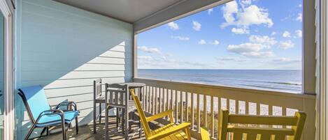 A covered outdoor area with a view of the ocean, featuring a blue chair, a yellow rocking chair, and a metal table.