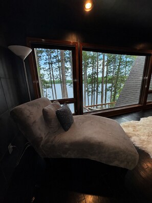 Een kamer met een groot raam, een chaise longue, een lamp en uitzicht op bomen en een wateroppervlak.
