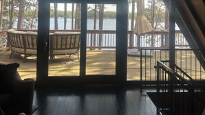 A spacious room with a large window overlooking a lake, furnished with a sofa and chairs, and a wooden deck outside.