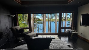 Une chambre avec une grande fenêtre donnant sur un lac, un lit, une chaise et un téléviseur fixé au mur.