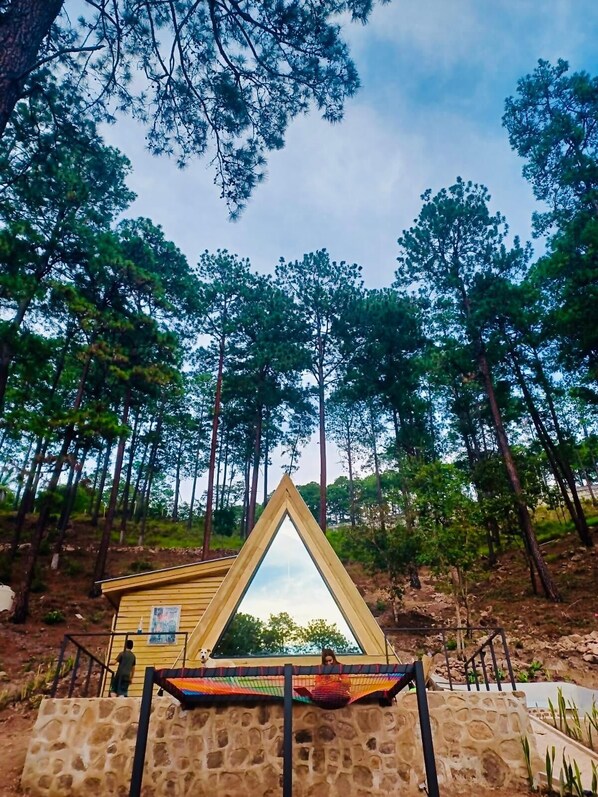 Estructura de madera triangular con una hamaca y un perro cerca, rodeada por un muro de piedra y árboles.