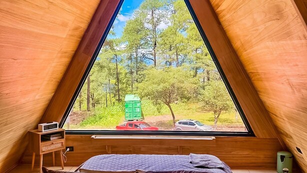A triangular wooden room with a bed, a small table, and a window offering a view of trees and a parked car.