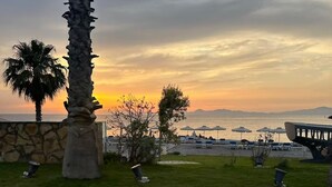 A coastal view with a sunset, palm trees, and a beach area with umbrellas and lounge chairs. - 4 minutes by car from the beach new modern apartment in Rhodes town (Rodos)