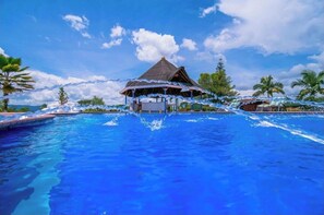 A swimming pool with a thatched-roof structure in the center, surrounded by palm trees and clear skies.