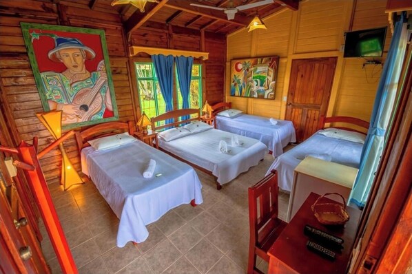 A wooden cabin with three beds, each with white linens and a towel. There are paintings on the walls, a television, and a wooden table with a basket and remote controls.