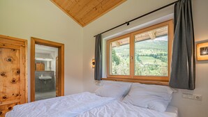 A bedroom with a wooden ceiling, a large window with a view of a green landscape, a bed with white linens, and a wooden wardrobe. - Cosy holiday flat with mountain views (Vorderkrimml)