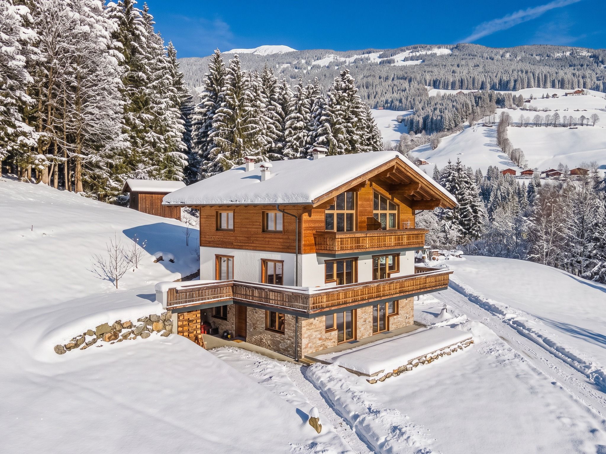 A two-story wooden chalet with a balcony, surrounded by snow-covered trees and a mountainous backdrop.