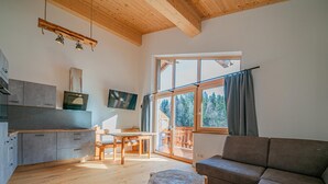 A modern living room with a wooden floor, a grey sofa, a wooden coffee table, a dining table with chairs, a kitchen with grey cabinets, a TV, and a large window with a view of trees. - Cosy holiday flat with mountain views (Vorderkrimml)