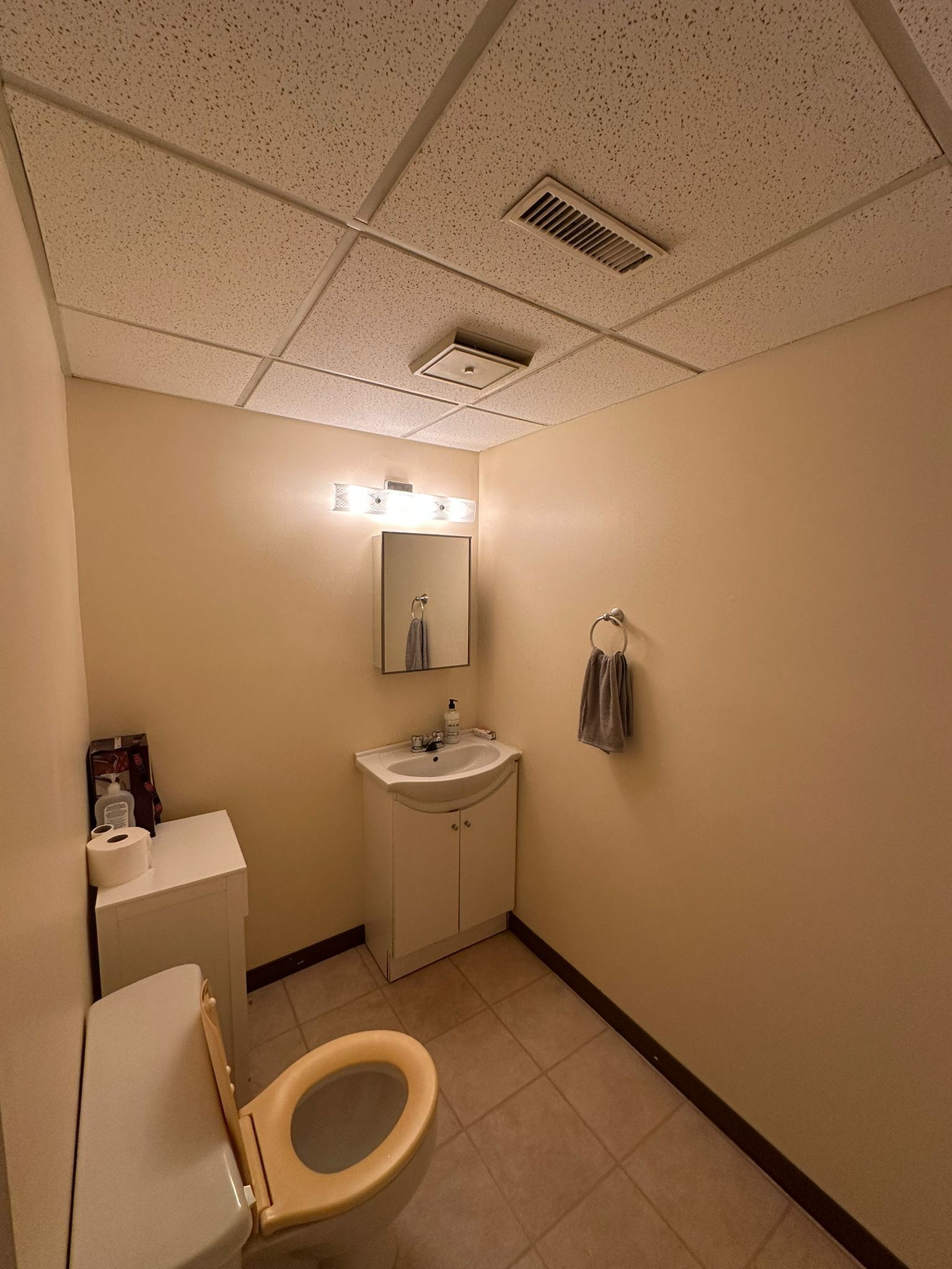 A small, plain bathroom with a toilet, sink, and mirror. There is a towel rack and a box of toilet paper on the counter.
