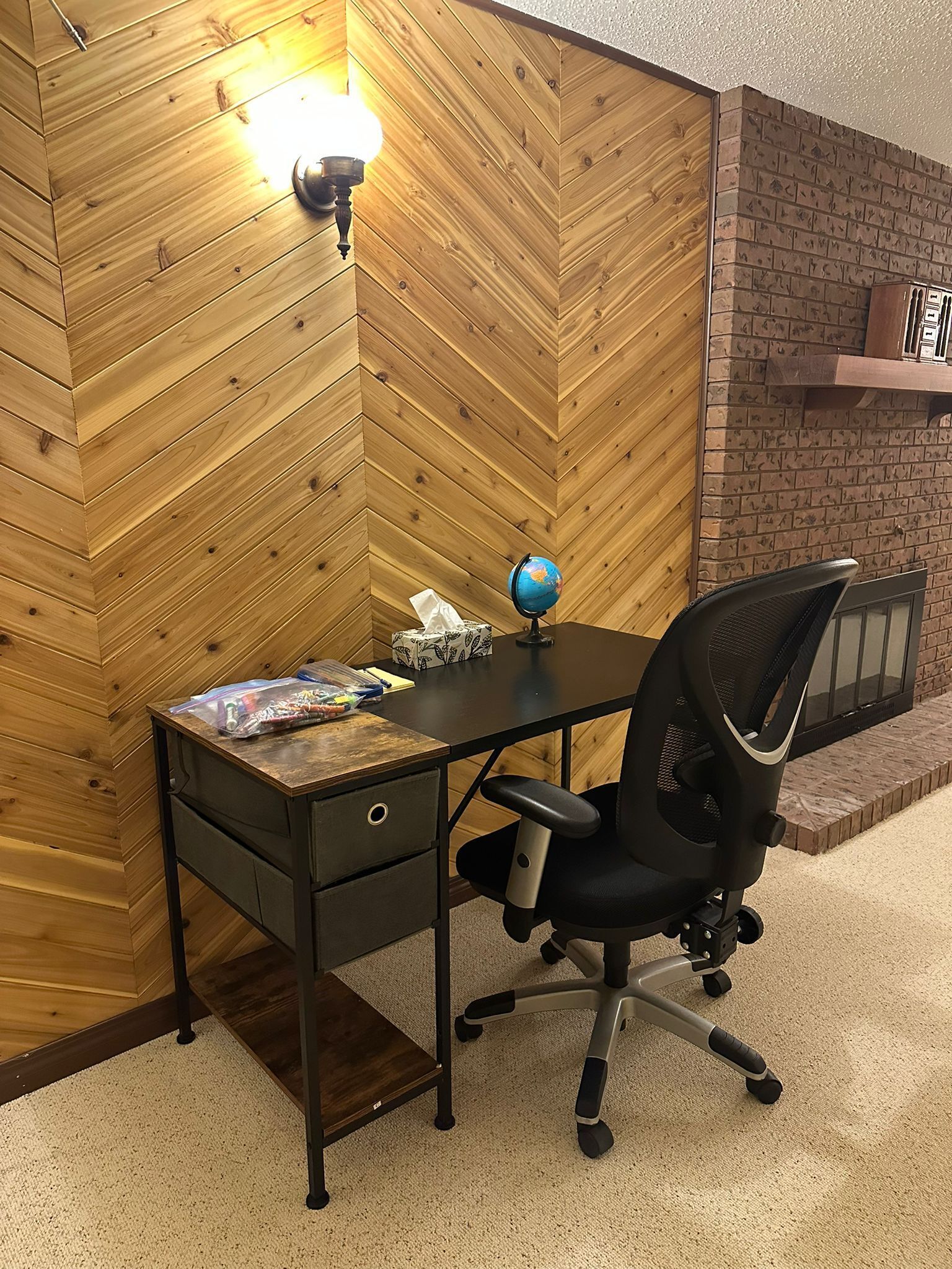 A wooden herringbone wall, a desk with a globe and tissues, and a black office chair.