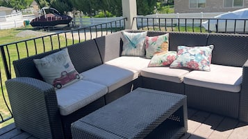 A covered outdoor seating area with wicker furniture, including a sofa and ottoman, and a boat in the background.