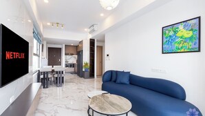 A modern living room with a blue sofa, a TV displaying 'NETFLIX', a dining area with chairs and a table, and a kitchen in the background.