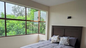 A bedroom with a large window, a bed with a grey bedspread, and a dark headboard. - Cozy apartment near the GNP stadium (Ciudad de México)
