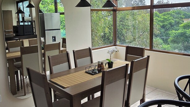 A dining area with a large table and chairs, a kitchen in the background, and a view of greenery through large windows. - Cozy apartment near the GNP stadium (Ciudad de México)