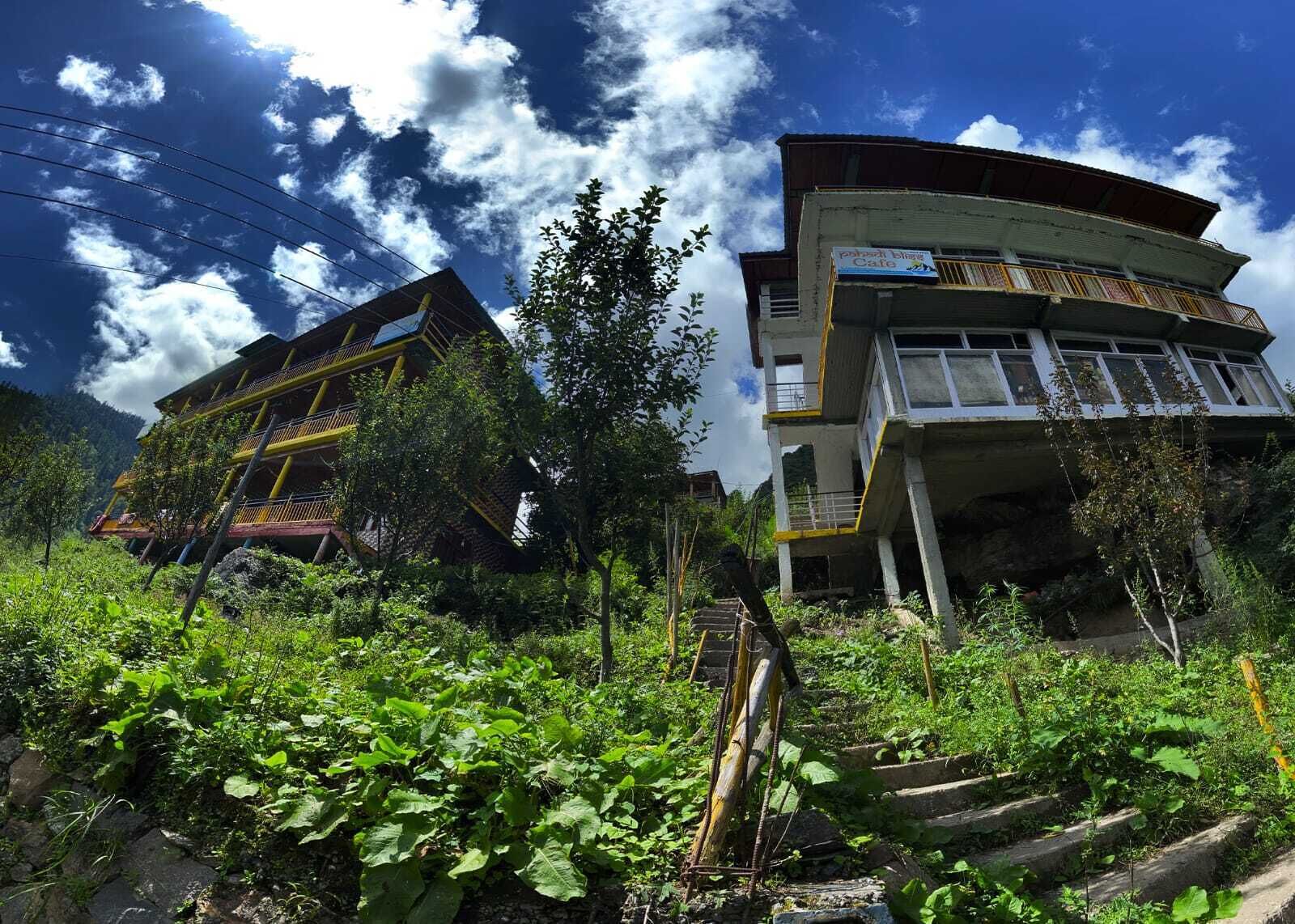 Edifício de dois andares com varanda no andar superior, cercado por vegetação exuberante e céu claro.