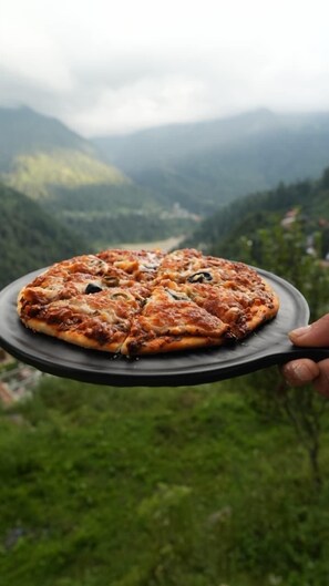 Une pizza sur une assiette noire, présentée devant un paysage pittoresque d’une vallée avec des montagnes en arrière-plan.