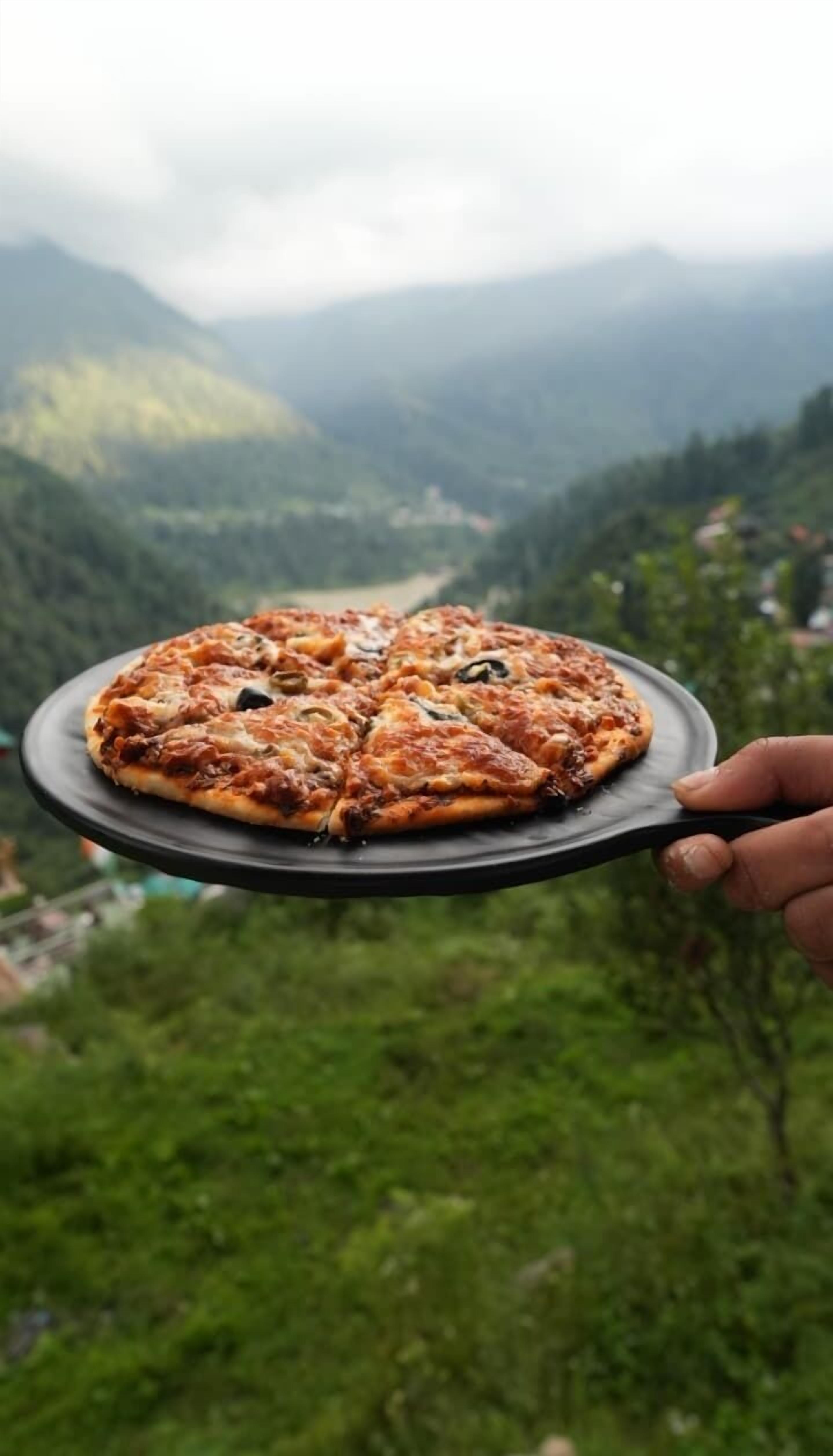 A hand holding a pizza on a plate with a mountainous landscape in the background.