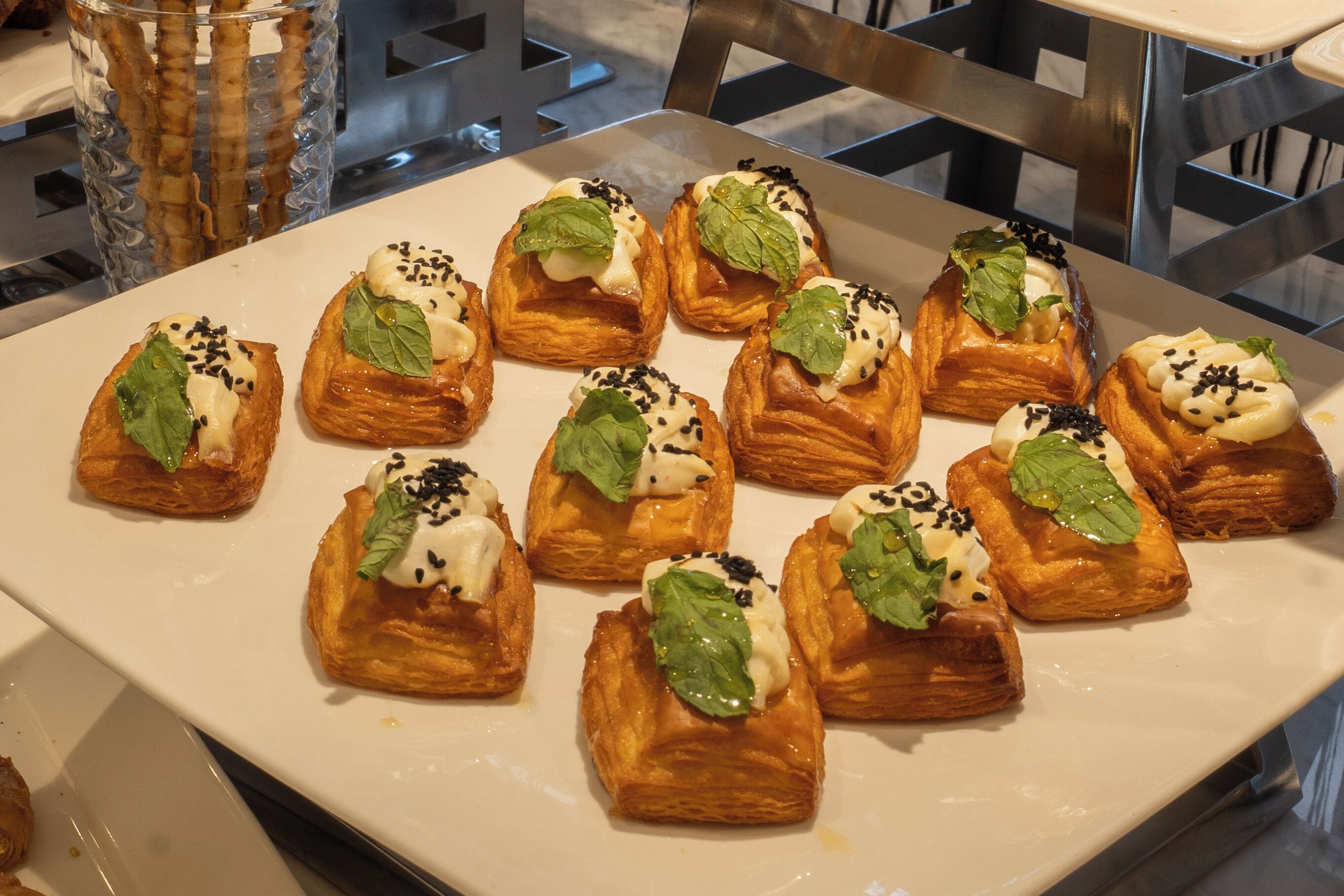 A white rectangular plate with small pastries topped with cream, black sesame seeds, and green leaves.