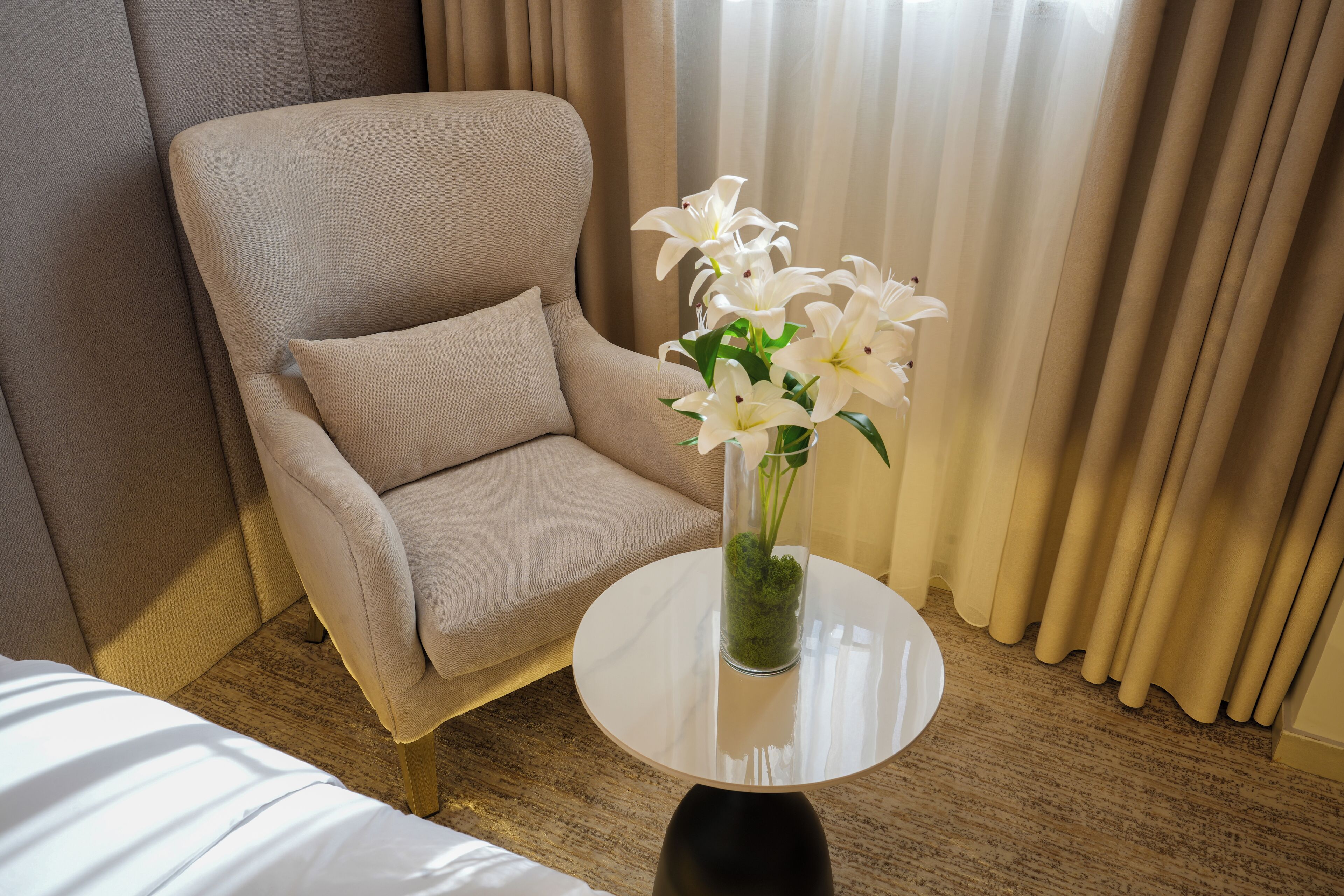 A beige armchair next to a round table with a clear vase containing white flowers.