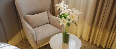 Un fauteuil beige est placé à côté d’une table ronde sur laquelle se trouve un vase transparent contenant des fleurs blanches.