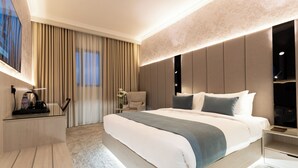 Quarto de hotel moderno com uma cama grande, uma mesinha de cabeceira com abajur, uma cadeira e uma TV.