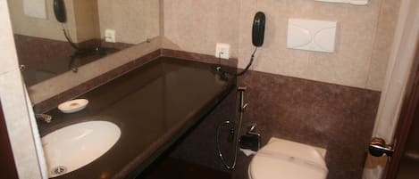 Une salle de bain avec un lavabo, des toilettes et un miroir. Il y a un porte-serviettes au-dessus des toilettes et un téléphone fixé au mur.