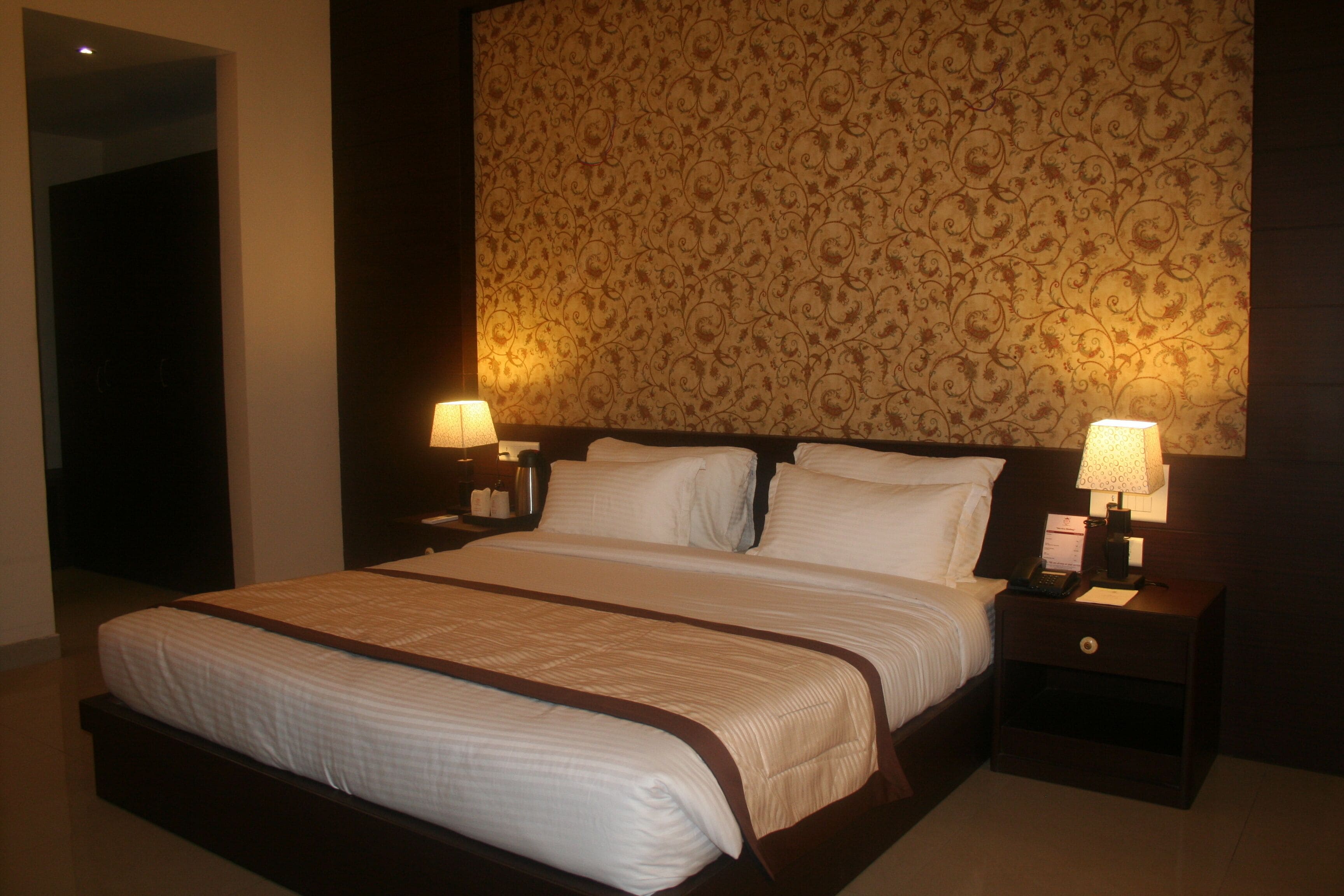 Une chambre d’hôtel avec un grand lit, deux tables de chevet avec des lampes, un papier peint à motifs et une tête de lit en bois foncé.