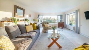 A spacious living room with a grey sofa, yellow cushions, a glass coffee table, and a view of the outdoors through large windows. - Fairway, Thorpeness, Aldeburgh, Suffolk (Thorpeness)