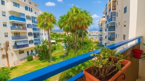 A balcony with a potted plant, overlooking a pool area with palm trees and a view of other buildings. - Apartamento Las Brisas (Estepona)