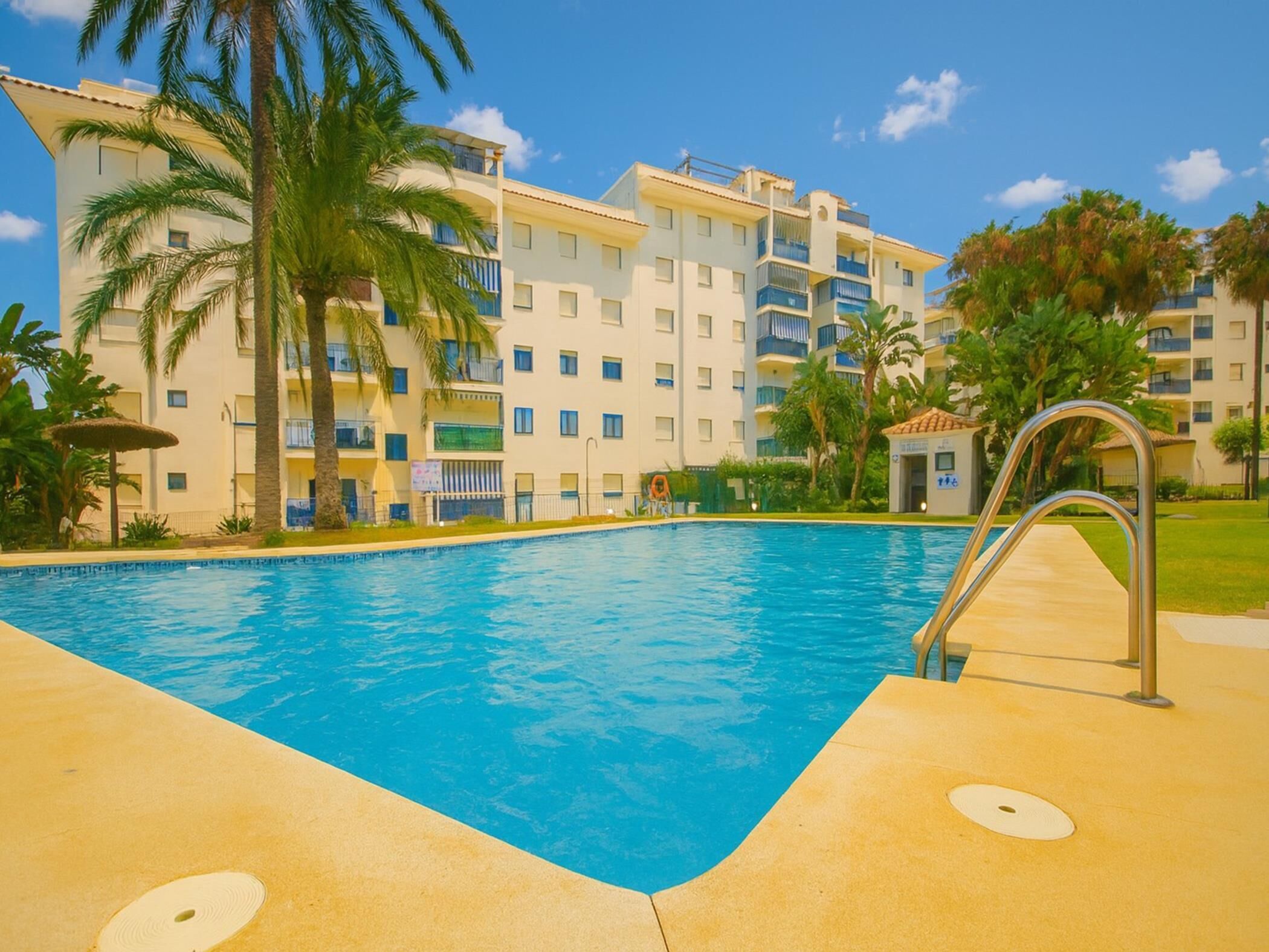 A swimming pool with a lifeguard stand, palm trees, and a multi-story building with balconies.