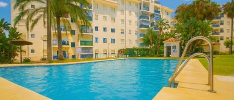 A swimming pool with a lifeguard stand, palm trees, and a multi-story building with balconies.