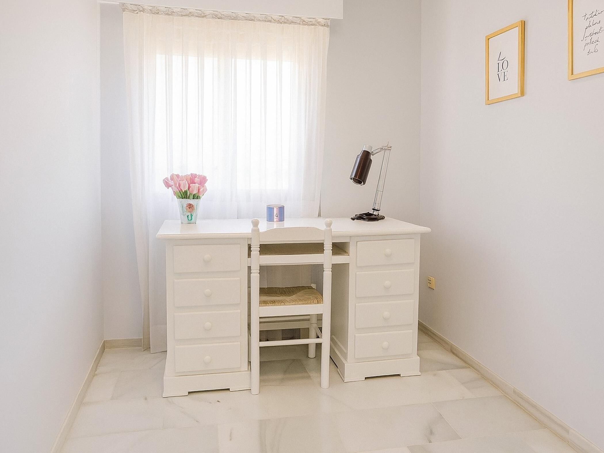 A white desk with drawers, a lamp, a vase with flowers, and a window with sheer curtains.