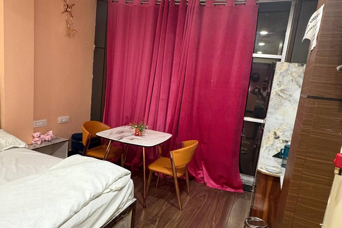 Habitación de hotel pequeña y funcional con cama, una mesita con sillas, ventilador de techo y ventana con cortinas.