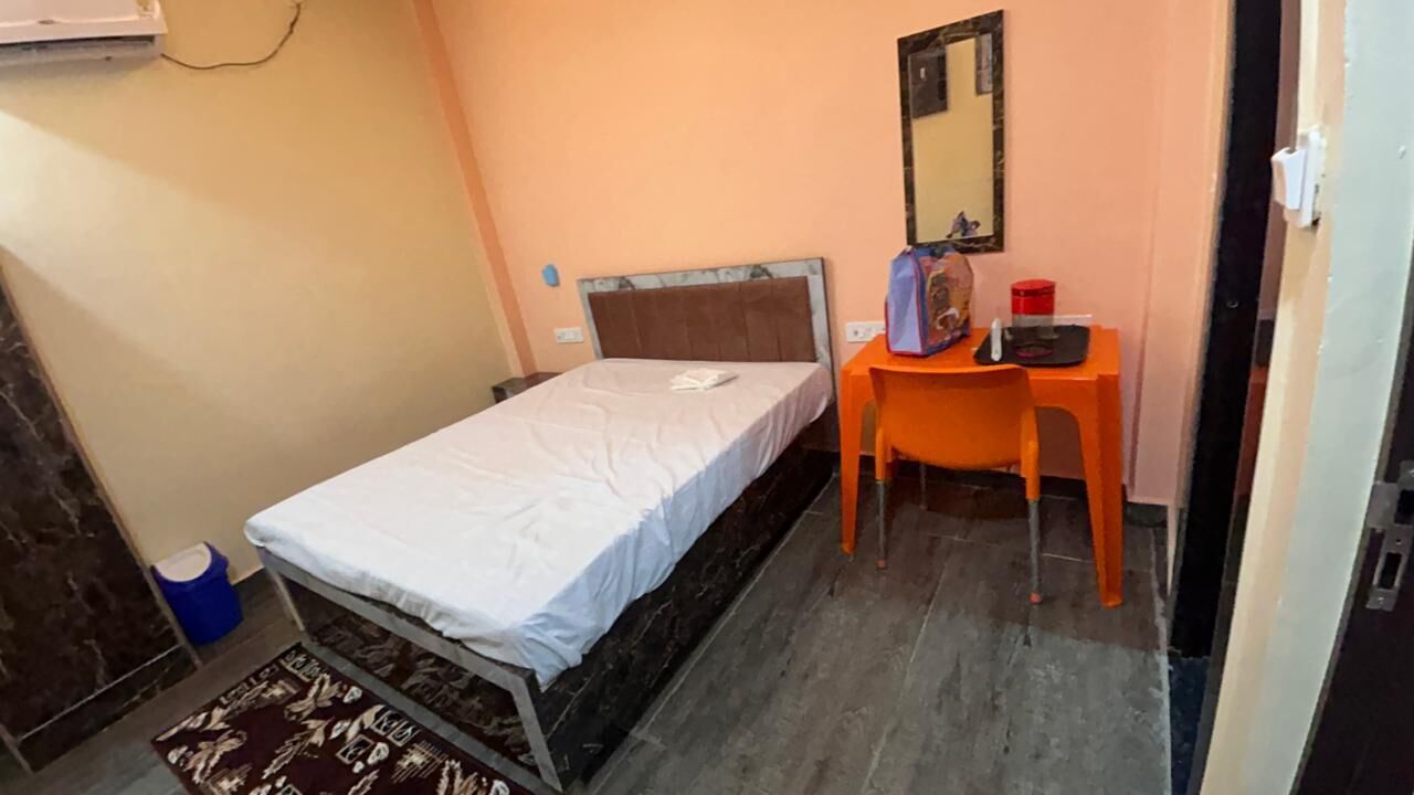 Una habitación pequeña con una cama individual, un escritorio y una silla. Hay un espejo en la pared y un cubo de basura cerca de la puerta.