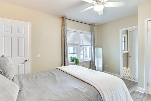 Un dormitorio con cama, ventilador de techo, ventana con persianas y puerta que da a otra habitación.