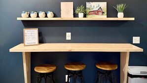 Un espace bar agrémenté de deux lampes suspendues, d’un comptoir en bois, de trois tabourets et d’étagères où sont disposés des objets décoratifs et un panneau d’accueil encadré.