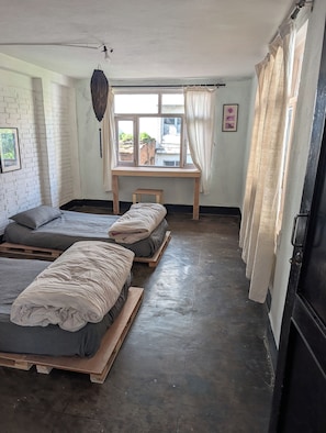 Habitació amb dos llits sobre palets de fusta, una taula petita, un llum penjant i una finestra amb cortines.