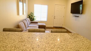 A living room with a large granite countertop, a beige sofa set, a flat-screen TV mounted on the wall, and a door in the background. - 2-bedroom guesthouse in Riverside. Centrally located near CBU,Parkview and 91fwy (Riverside)