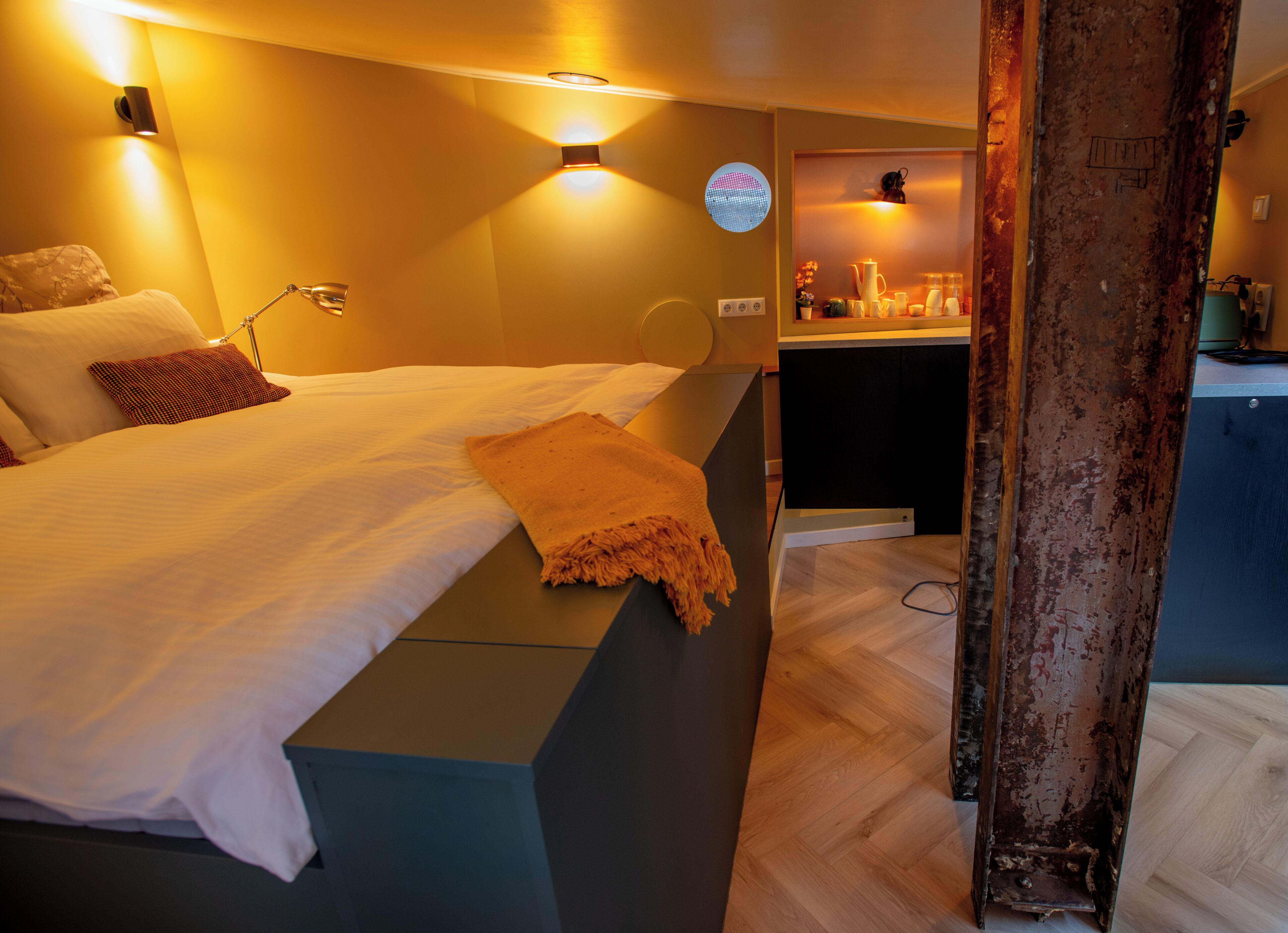 Een hotelkamer met een bed, nachtkastje, lamp en uitzicht op de keuken.