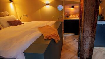 Een hotelkamer met een bed, nachtkastje, lamp en uitzicht op de keuken.