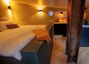 A hotel room with a bed, bedside table, lamp, and a view of the kitchen.