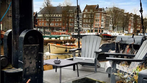 Un pont de bateau avec des chaises et une table, une plante en pot, et une vue sur un canal avec des bâtiments en arrière-plan.