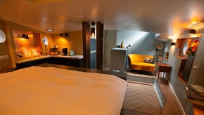 Une chambre d’hôtel avec un grand lit, un coin salon comprenant un fauteuil et une petite table, ainsi qu’un coin cuisine.