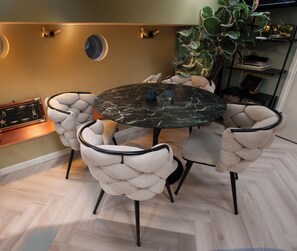 A dining area with a round table, four upholstered chairs, a potted plant, and a wall-mounted shelf with books.