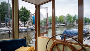 Una habitación con un ventanal grande con vistas a un canal con barcos, un sofá azul y una rueda de timón de madera.