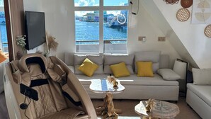 A modern living room with a massage chair, a sofa with yellow cushions, a coffee table, and a view of the water through large windows. - Atoll Villa (Hulhumalé)