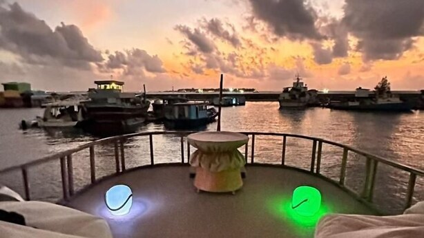 A deck with a round table and two chairs, overlooking a body of water with boats and a sunset. - Atoll Villa (Hulhumalé)