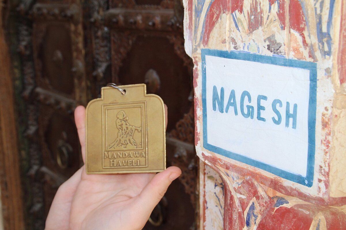 A hand holding a brass keychain with "MANDAWA HAVELI" engraved, and a wall with a sign that reads "NAGESH".