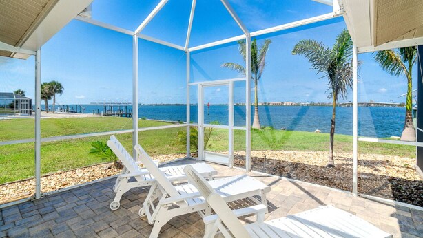 A covered outdoor seating area with white chairs, a view of the sea, and a palm tree.